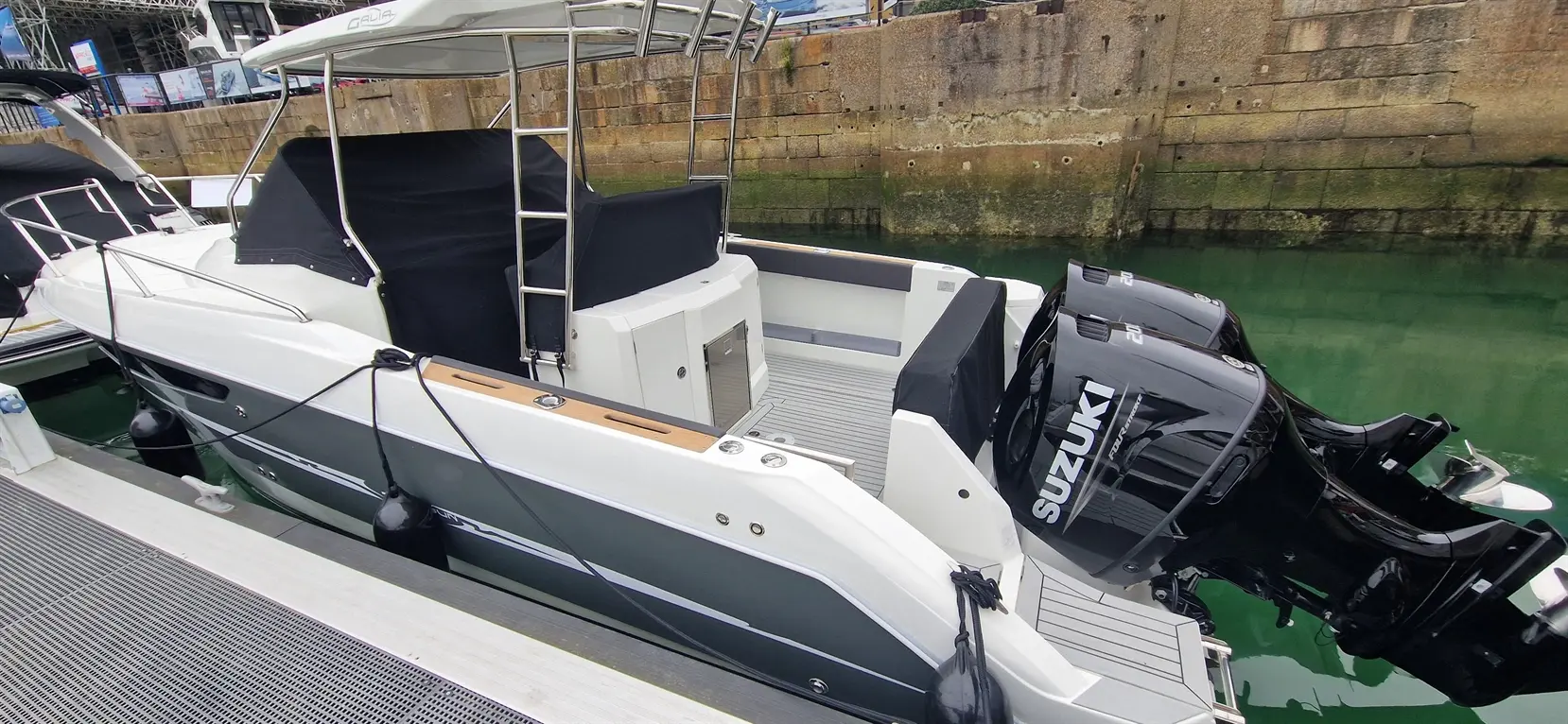 A sleek motorboat docked near a stone wall, featuring two Suzuki outboard motors and a spacious deck with gray flooring.